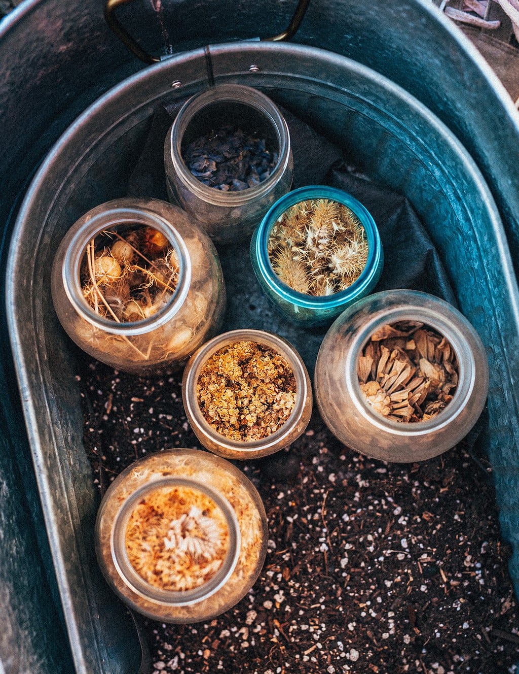 A galvanized steel tub with soil, holding jars with different types of seeds.